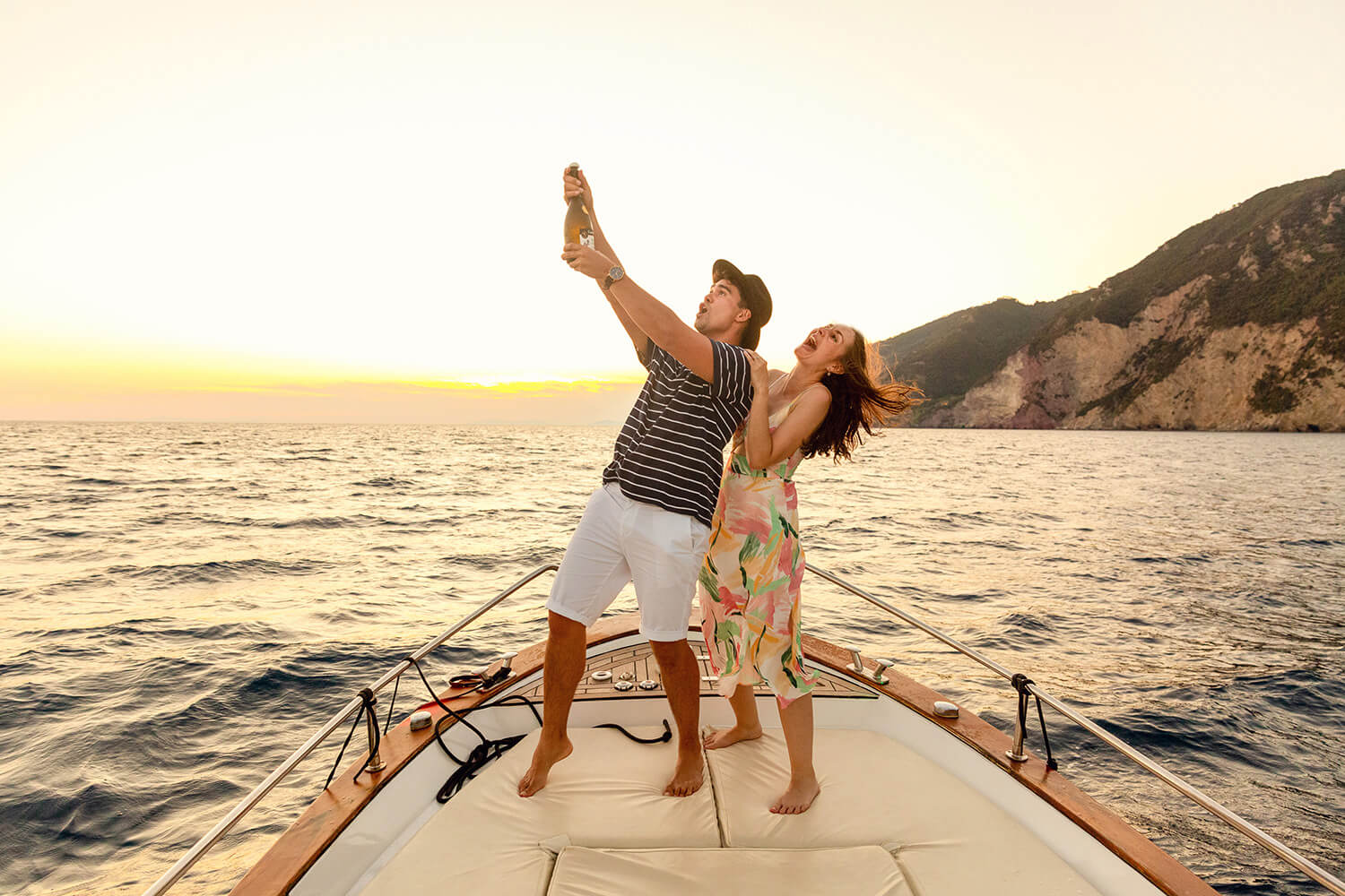 Couple popping champagne on a boat at sunset to celebrate their engagement after a marriage proposal